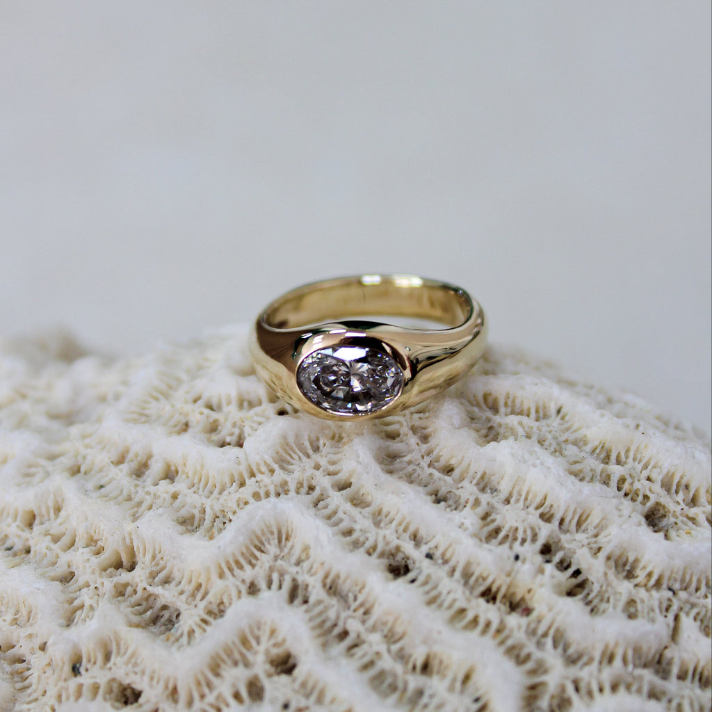 A gold ring with an oval diamond gemstone resting on a textured beige coral shell against a light gray background.
