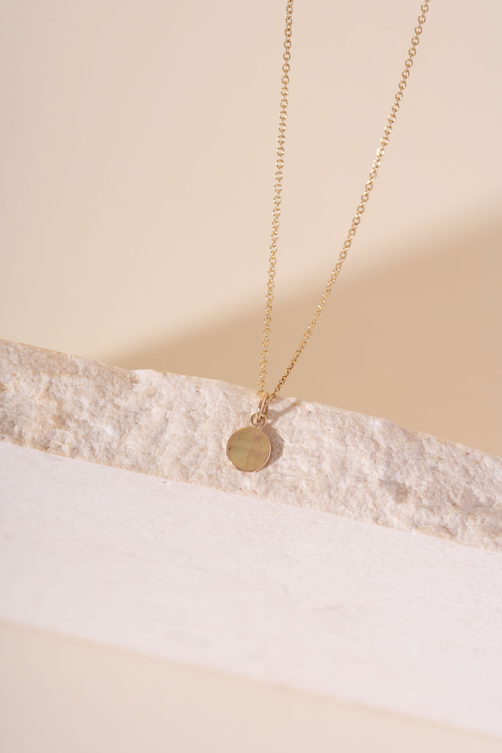 A delicate gold chain necklace with a small, flat circular disc pendant, resting on a textured white stone slab against a warm beige background.