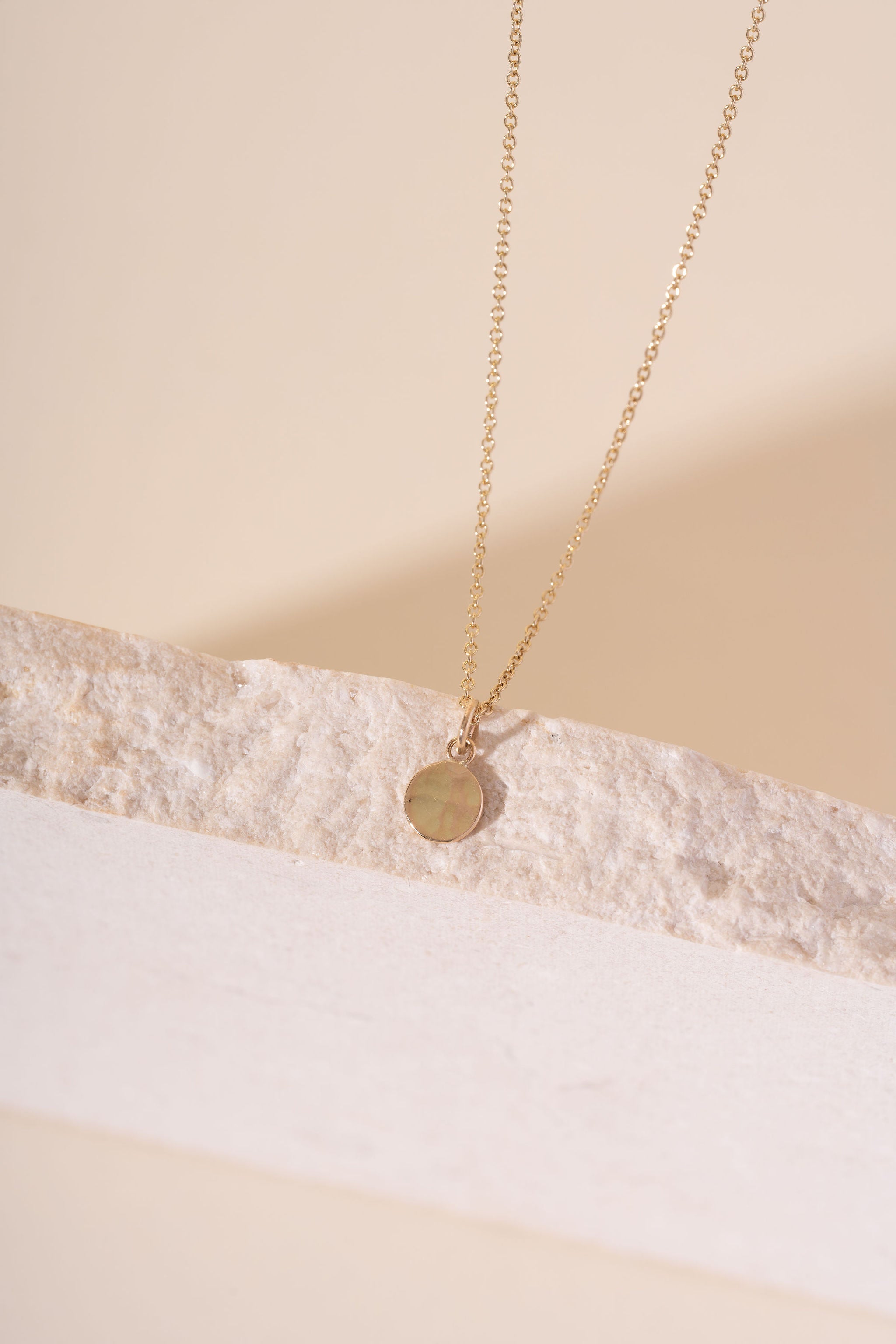 A delicate gold chain necklace with a small, flat circular disc pendant, resting on a textured white stone slab against a warm beige background.