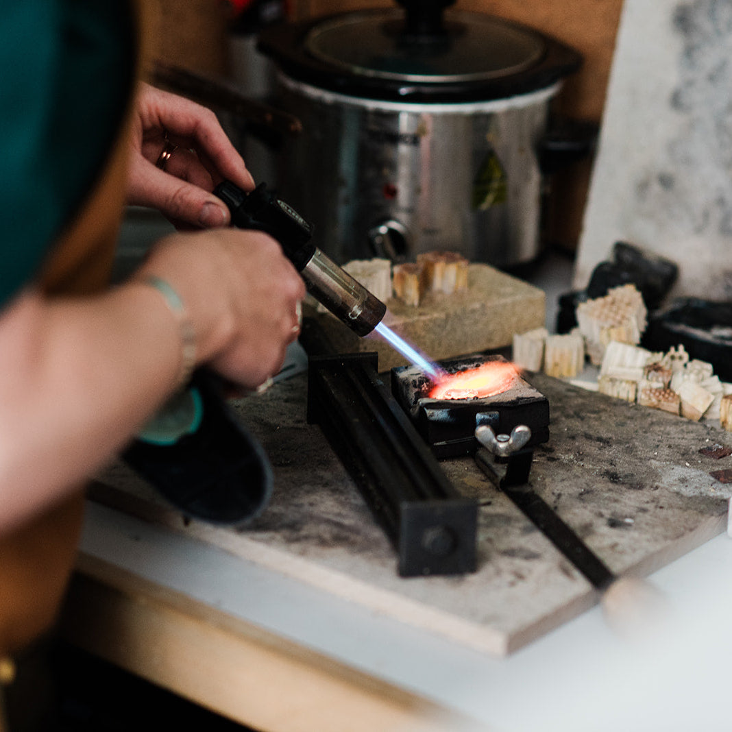 Person using a torch on a piece of jewelry in a workshop setting.